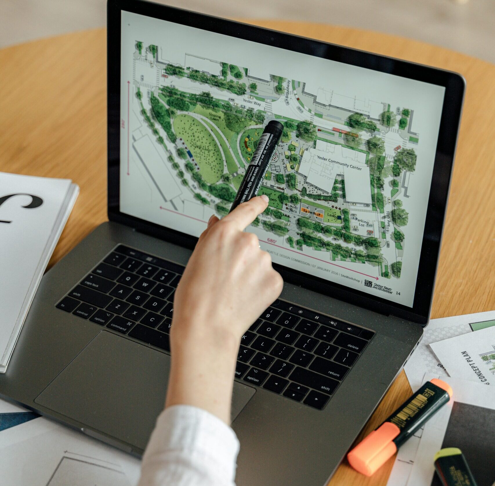 An architect reviews landscape design plans on a laptop, using highlighters and documents at a wooden desk.